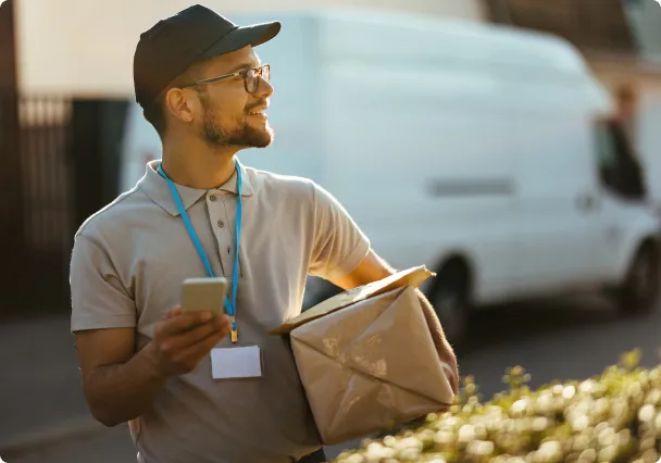 Un livreur consulte son téléphone, un colis sous le bras, sa camionnette derrière lui