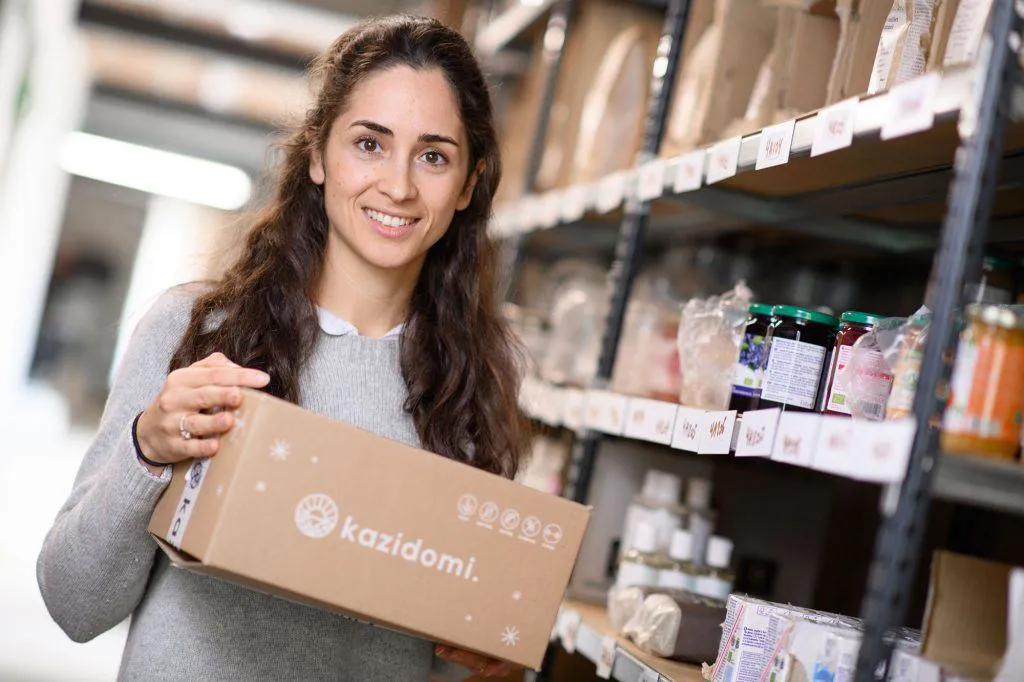 Une employée de Kazidomi tient un carton de la marque dans une allée de l’entrepôt, devant des étagères de bocaux