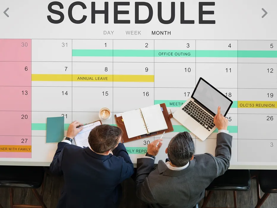 Deux personnes en costume, vues de dessus, travaillent à une table posée sur un grand calendrier mensuel intitulé SCHEDULE